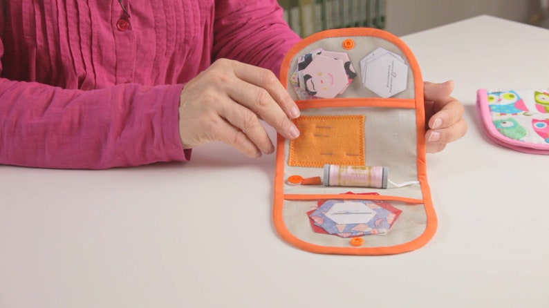 Op de afbeelding: Een open naaiset van beige stof met een oranje rand. De set bevat een spoel garen, naalden en stofresten.