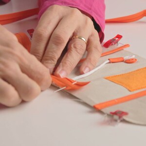 Op de afbeelding: Een persoon naait een beige stoffen tasje met een oranje rand. Het tasje heeft een klein oranje vakje en een wit koord. Het tasje wordt bij elkaar gehouden met rode clips.