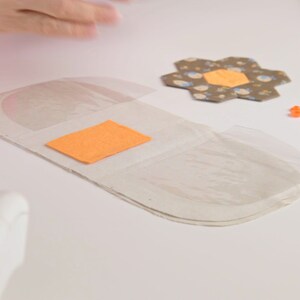 Op de afbeelding: Een wit stoffen kussen met een transparante plastic overlay en een oranje vilt vierkant. Het kussen ligt op een wit oppervlak met een stoffen bloem, oranje knopen en een wit touwtje.