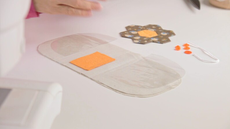 Op de afbeelding: Een wit stoffen kussen met een transparante plastic overlay en een oranje vilt vierkant. Het kussen ligt op een wit oppervlak met een stoffen bloem, oranje knopen en een wit touwtje.