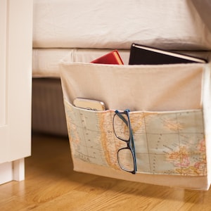 Puede incluir: Un organizador de cama de tela beige con un bolsillo estampado con un mapa. El organizador cuelga del marco de la cama y contiene un libro rojo, un libro negro, un teléfono y gafas. Las gafas tienen monturas azules.
