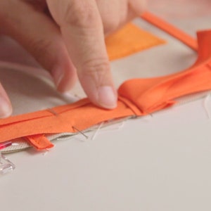 Op de afbeelding: Een close-up van een hand die oranje stof op een wit oppervlak vastpint. De stof is gevouwen en vastgezet met kleine clips.