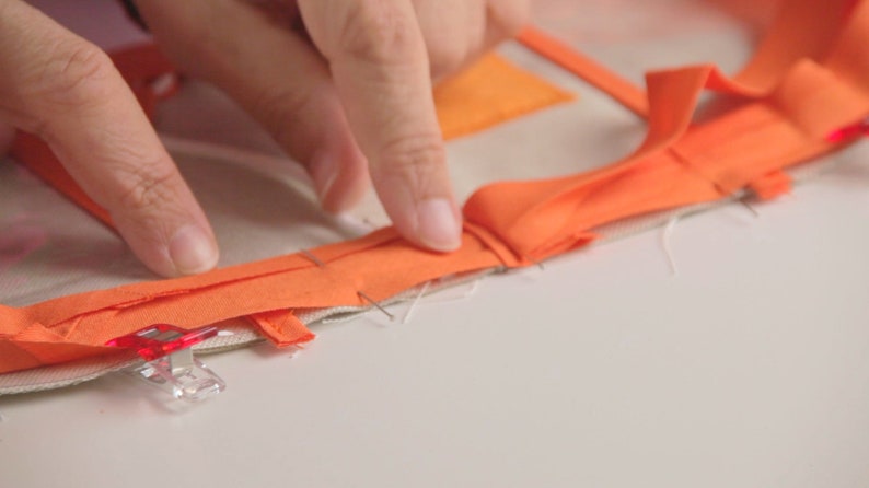 Op de afbeelding: Een close-up van een hand die oranje stof op een wit oppervlak vastpint. De stof is gevouwen en vastgezet met kleine clips.