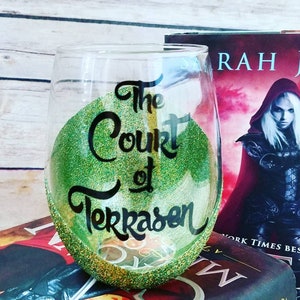 May include: A green and gold glitter wine glass with the text "The Court of Terrasen" written in black. The glass is sitting on top of a stack of books with the title "Queen of Midnight" visible.