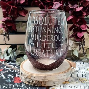 May include: A clear wine glass with a glittery burgundy finish. The glass has the text "Absolutely Stunning, Murderous Little Creature" printed on it.
