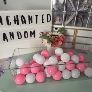 以下が含まれることがあります： 白とピンクのボールが入った透明なプラスチック容器。背景には「ENCHANTED FANDOM」という文字が入ったライトアップサインと、小さな花のブーケがあります。シーンは明るい緑色の表面に設定されています。