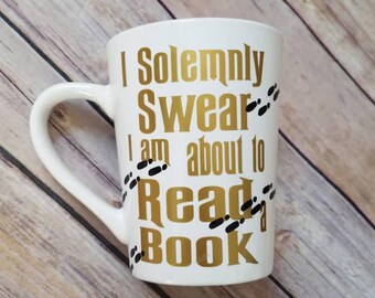 May include: White ceramic mug with a gold inscription that reads "I Solemnly Swear I Am About To Read A Book" with black footprints around the text.