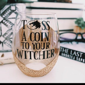 May include: A clear glass wine glass with a gold glitter rim. The glass has a black vinyl decal with the text "Toss a coin to your Witcher" and a wolf head graphic.