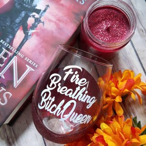 May include: A red wine glass with glitter on the rim and the text "Fire Breathing Bitch Queen" in white lettering. The glass is sitting on a wooden surface with a book and a candle in the background.