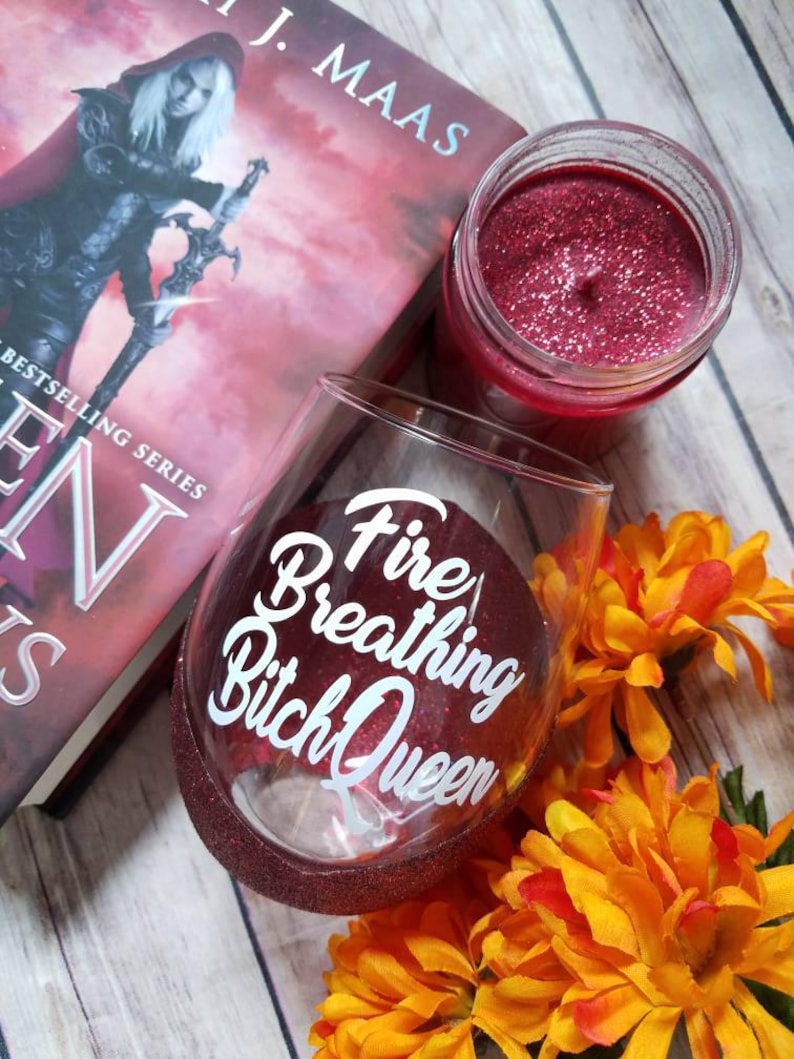 May include: A red wine glass with glitter on the rim and the text "Fire Breathing Bitch Queen" in white lettering. The glass is sitting on a wooden surface with a book and a candle in the background.