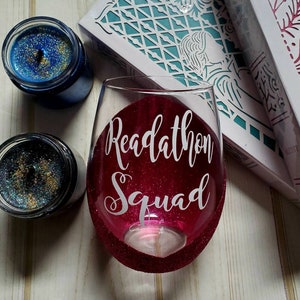 May include: A pink wine glass with glitter on the bottom and sides. The glass has a white vinyl decal that says "Readathon Squad".