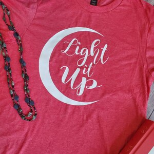 K&ouml;nnte beinhalten: Ein rotes T-Shirt mit dem Schriftzug "Light it Up" in Wei&szlig; in einem wei&szlig;en Halbmond. Eine Perlenkette und ein karierter Hut sind im Hintergrund zu sehen.