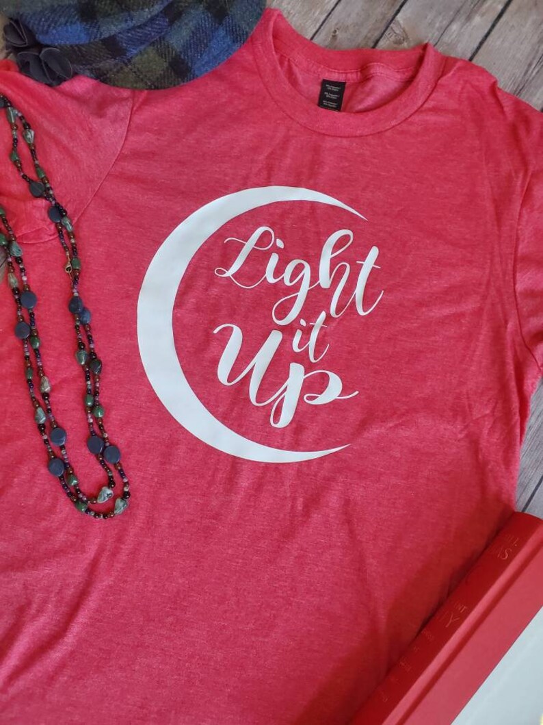 K&ouml;nnte beinhalten: Ein rotes T-Shirt mit dem Schriftzug "Light it Up" in Wei&szlig; in einem wei&szlig;en Halbmond. Eine Perlenkette und ein karierter Hut sind im Hintergrund zu sehen.