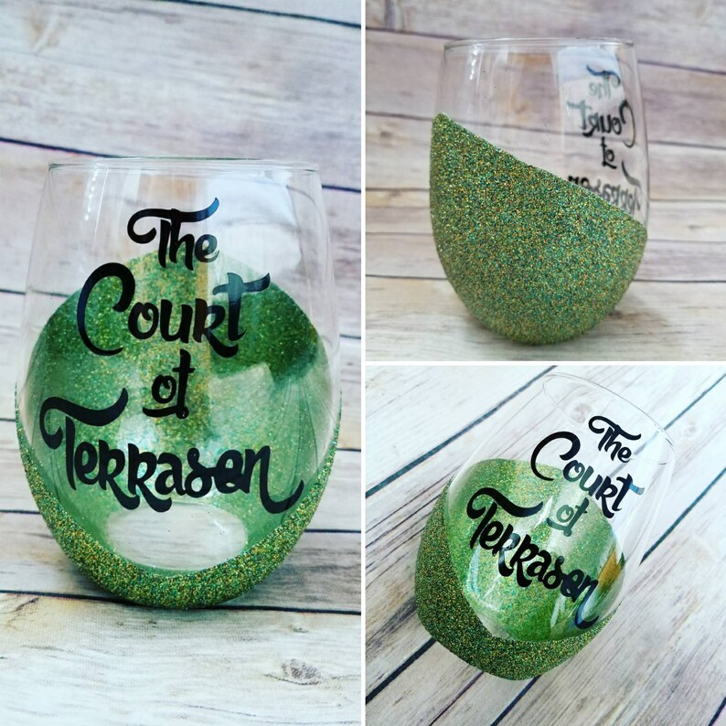 May include: A clear glass wine glass with a green glitter rim and the text "The Court of Terrasen" written in black on the side.