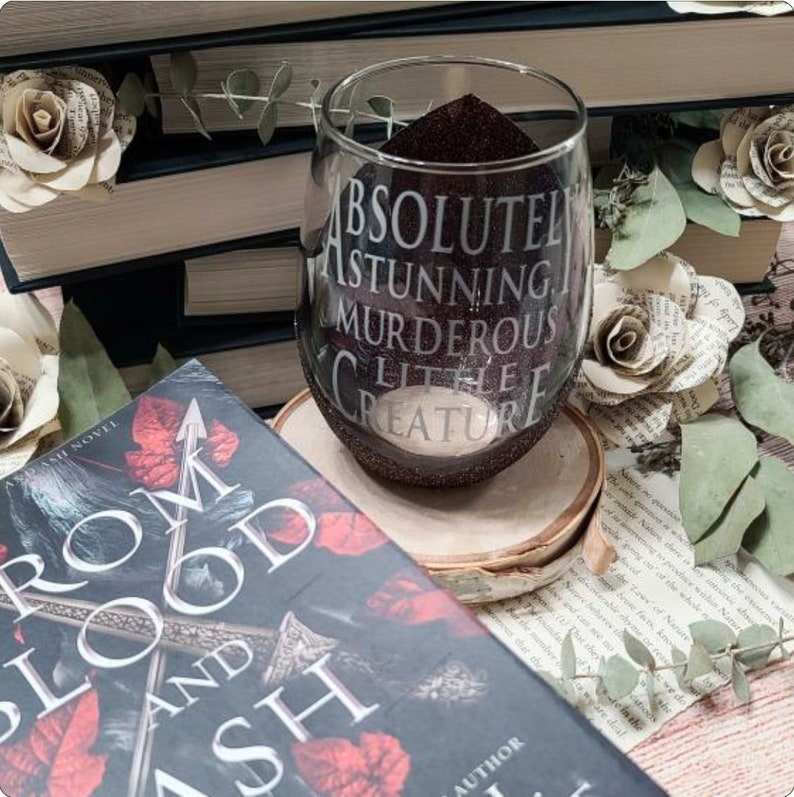 May include: A dark red wine glass with glitter on the outside. The glass has the text "ABSOLUTELY STUNNING MURDEROUS LITTLE CREATURE" printed on it. The glass is sitting on a wooden coaster. A book titled "From Blood and Ash" is in the background.