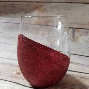 May include: A clear glass wine glass with a red glitter design on the bottom half. The glass has the text "It Goes" written on the side.