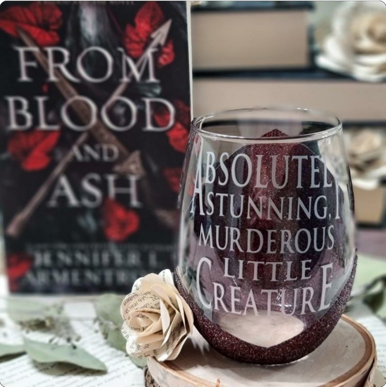 May include: A wine glass with a burgundy glitter finish and the text "ABSOLUTE STUNNING, MURDEROUS LITTLE CREATURE" etched on the side. The glass is sitting on a wooden surface next to a book titled "From Blood and Ash" by Jennifer L. Armentrout.