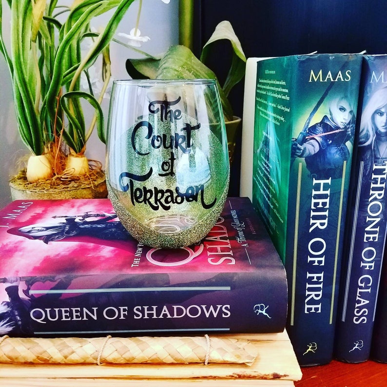 May include: A clear glass wine glass with gold glitter and black lettering that reads "The Court of Terrasen". The glass is sitting on top of a stack of three books. The books are all by Sarah J. Maas and are titled "Throne of Glass", "Heir of Fire", and "Queen of Shadows".