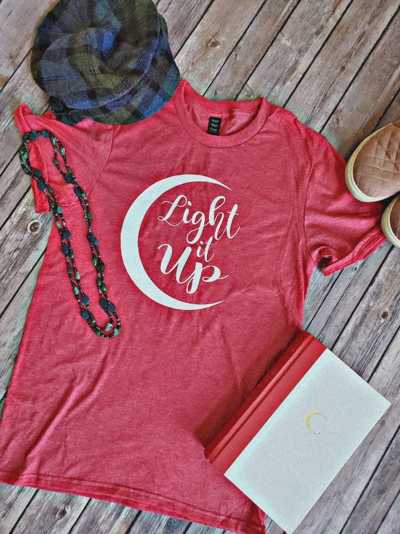 K&ouml;nnte beinhalten: Ein rotes T-Shirt mit dem wei&szlig;en Schriftzug "Light it Up" und einem Mond-Design. Das Bild zeigt auch einen karierten Hut, eine Perlenkette, ein Buch und Turnschuhe. Die Artikel sind auf einer Holzoberfl&auml;che angeordnet.