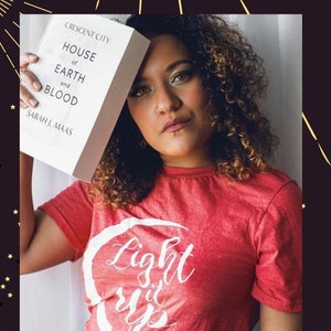 K&ouml;nnte beinhalten: Eine Person h&auml;lt ein Exemplar von "House of Earth and Blood" von Sarah J. Maas. Sie tr&auml;gt ein rotes T-Shirt mit dem wei&szlig;en Schriftzug "Light it Up". Das Bild hat eine warme, einladende Farbpalette.