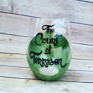 May include: A clear glass wine glass with a green glitter rim and green glitter inside. The glass has black lettering that reads "The Court of Terrasen".