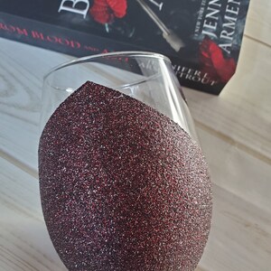 May include: A clear glass wine glass with a red glitter coating on the bottom half. The glass is sitting on a wooden surface. The book "From Blood and Ash" by Jennifer Armentrout is visible in the background.