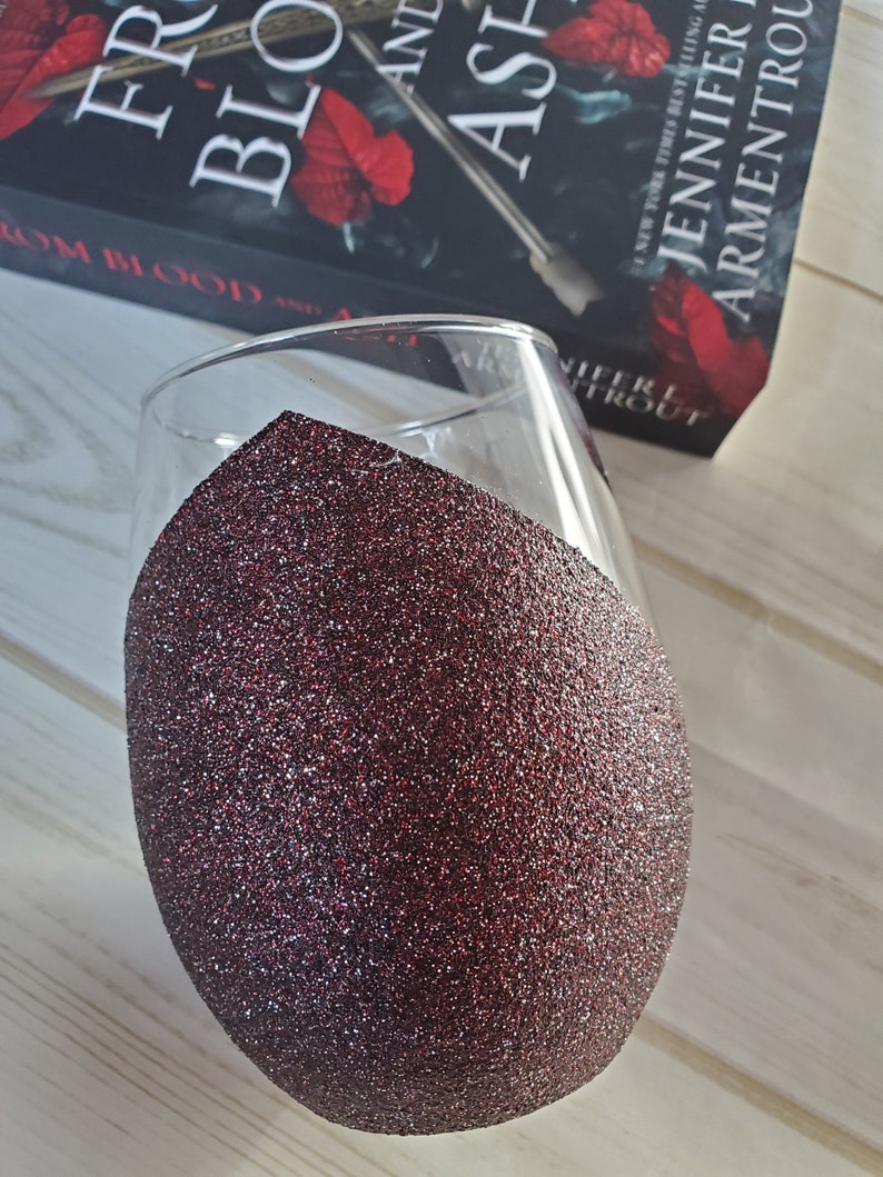 May include: A clear glass wine glass with a red glitter coating on the bottom half. The glass is sitting on a wooden surface. The book "From Blood and Ash" by Jennifer Armentrout is visible in the background.