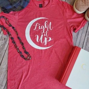 K&ouml;nnte beinhalten: Ein rotes T-Shirt mit dem wei&szlig;en Schriftzug "Light it Up" in einem Mond-Design. Das Bild zeigt einen Hut, eine Halskette, Schuhe und ein Buch mit einem goldenen Mond auf dem Einband.