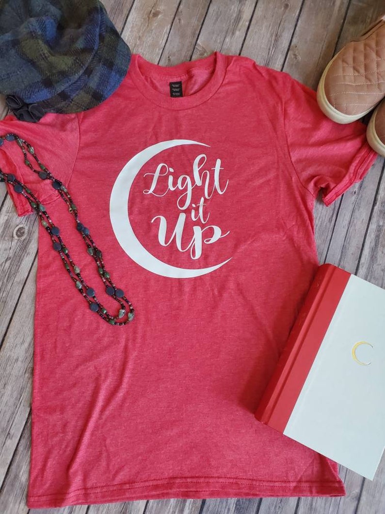 K&ouml;nnte beinhalten: Ein rotes T-Shirt mit dem wei&szlig;en Schriftzug "Light it Up" in einem Mond-Design. Das Bild zeigt einen Hut, eine Halskette, Schuhe und ein Buch mit einem goldenen Mond auf dem Einband.