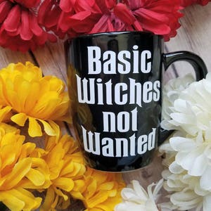 May include: Black coffee mug with white text that reads "Basic Witches Not Wanted". The mug is surrounded by yellow and white flowers.