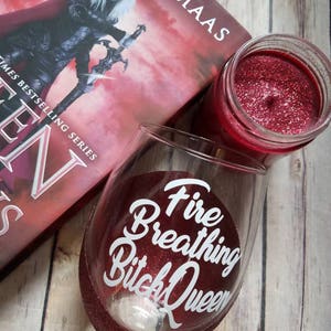 May include: A red glitter wine glass with the text "Fire Breathing Bitch Queen" in white. The glass is sitting on a wooden surface next to a book and a red candle.