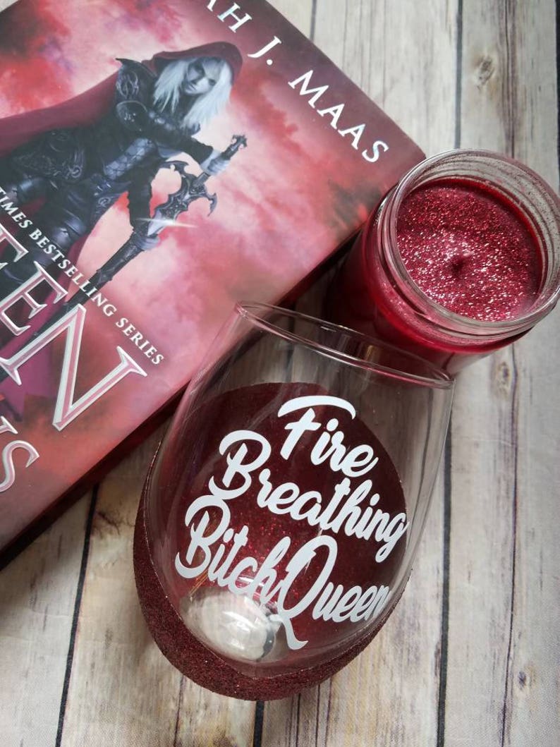 May include: A red glitter wine glass with the text "Fire Breathing Bitch Queen" in white. The glass is sitting on a wooden surface next to a book and a red candle.