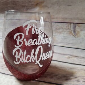 May include: A clear glass wine glass with a red glitter rim. The glass has the text "Fire Breathing Bitch Queen" written on it in white.