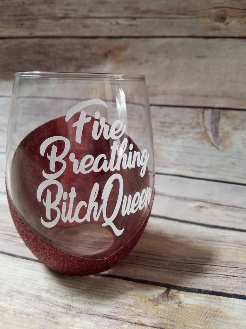 May include: A clear glass wine glass with a red glitter rim. The glass has the text "Fire Breathing Bitch Queen" written on it in white.