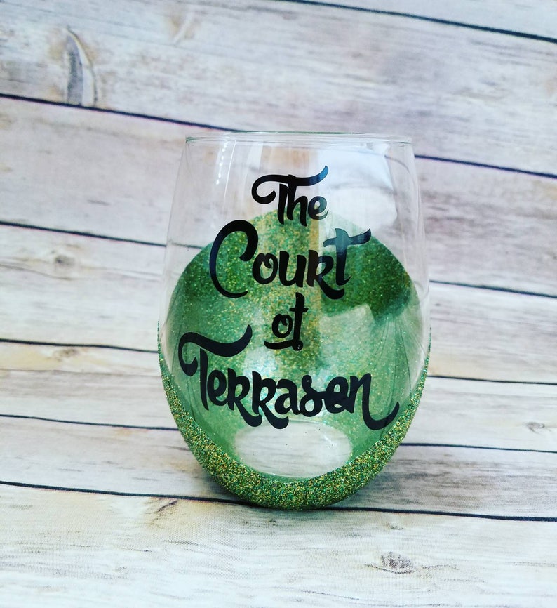 May include: A clear glass wine glass with a green glitter rim and green glitter inside. The glass has black lettering that reads "The Court of Terrasen".