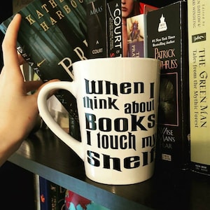 May include: White ceramic mug with black text that reads "When I think about BOOKS I touch my shelf". The mug is on a bookshelf with other books.