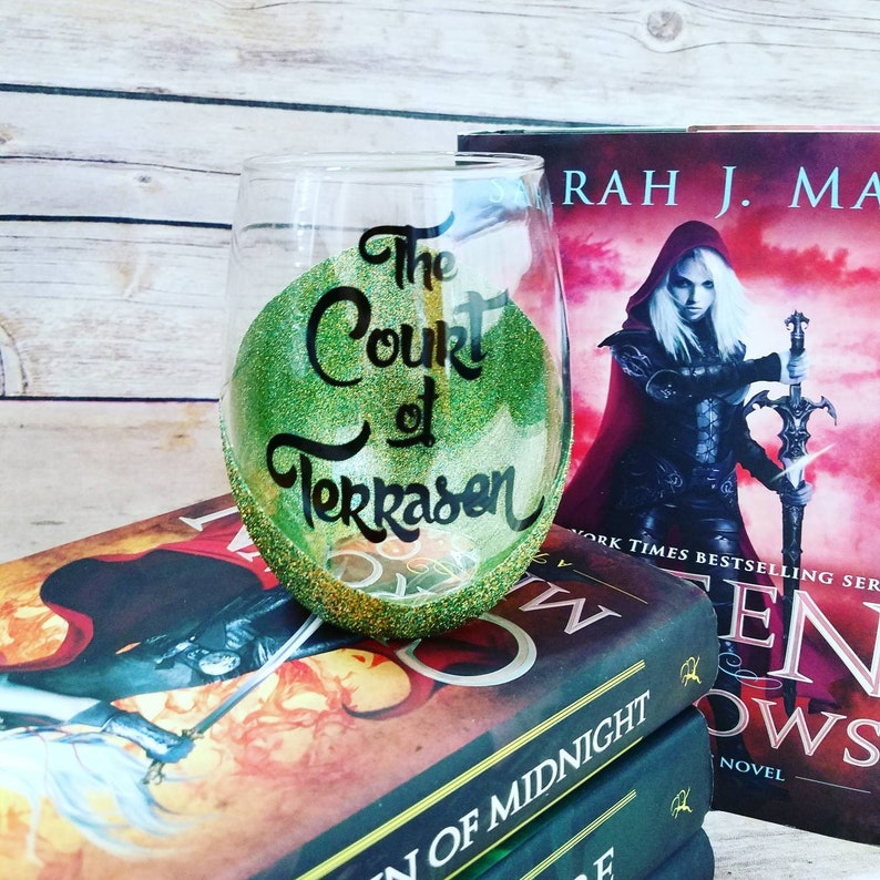 May include: A green and gold glitter wine glass with the text "The Court of Terrasen" written in black. The glass is sitting on top of a stack of books with the title "Queen of Midnight" visible.