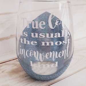 Könnte beinhalten: Ein stieloses Weinglas mit blauem Glitzerboden und blauem Glitzerdesign im Inneren. Das Glas trägt den weißen Schriftzug "True love is usually the most inconvenient kind".