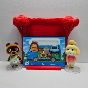May include: Two small animal figurines stand beside a rectangular trading card featuring a blue camper van and a lion character. The card is held by a red plastic stand shaped like a tree. The card displays the number 15 and the name Rex.