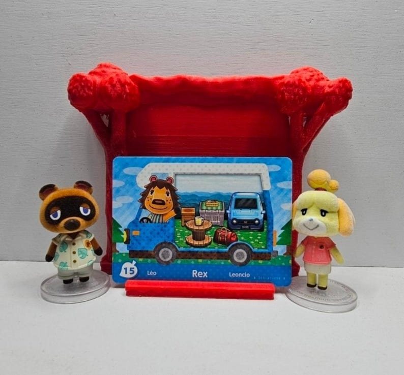 May include: Two small animal figurines stand beside a rectangular trading card featuring a blue camper van and a lion character. The card is held by a red plastic stand shaped like a tree. The card displays the number 15 and the name Rex.
