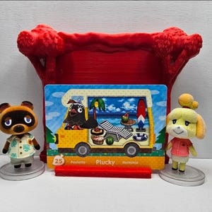 May include: A collectible Animal Crossing trading card featuring Plucky in a yellow camper van, displayed in a red plastic stand between two small figurines of Tom Nook and Isabelle. The card shows the number 25 and the name Plucky in white text.