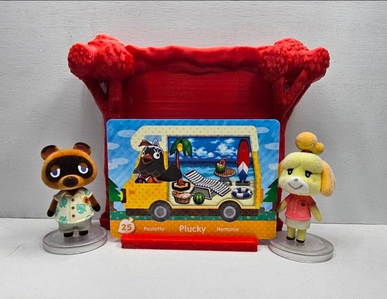 May include: A collectible Animal Crossing trading card featuring Plucky in a yellow camper van, displayed in a red plastic stand between two small figurines of Tom Nook and Isabelle. The card shows the number 25 and the name Plucky in white text.