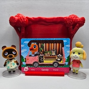 May include: A red 3D-printed display stand holding a rectangular Animal Crossing trading card featuring the character Sandy in a pink camper van. The card is flanked by two small figurines: a brown raccoon character and a yellow dog character.