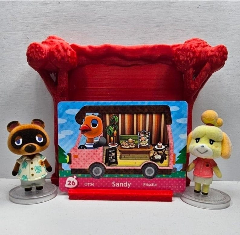 May include: A red 3D-printed display stand holding a rectangular Animal Crossing trading card featuring the character Sandy in a pink camper van. The card is flanked by two small figurines: a brown raccoon character and a yellow dog character.