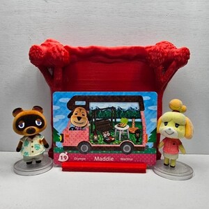 May include: Two small animal figurines stand on circular bases flanking a rectangular trading card. The card features a pink camper van with the name Maddie printed in the center. A red 3D-printed tree-shaped stand holds the card upright.