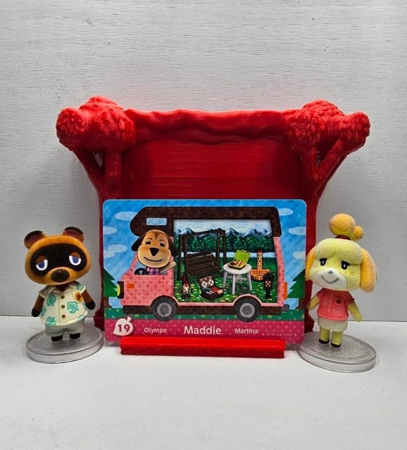 May include: Two small animal figurines stand on circular bases flanking a rectangular trading card. The card features a pink camper van with the name Maddie printed in the center. A red 3D-printed tree-shaped stand holds the card upright.