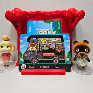 May include: Two small animal figurines stand beside a red plastic display stand holding an Animal Crossing Claude character card. The card features a brown rabbit character inside a camper van, with the text 27, Claude, and Pablo printed below.