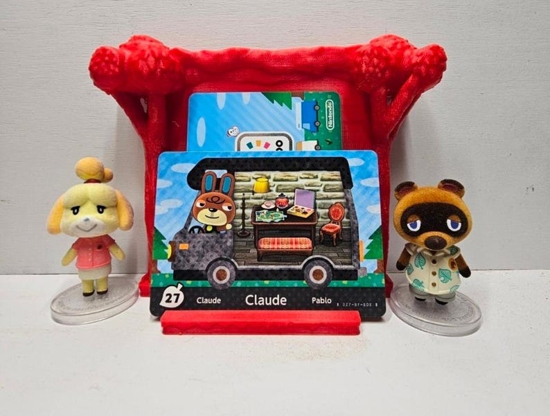 May include: Two small animal figurines stand beside a red plastic display stand holding an Animal Crossing Claude character card. The card features a brown rabbit character inside a camper van, with the text 27, Claude, and Pablo printed below.