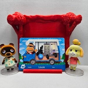 May include: A red plastic display stand holding a rectangular Animal Crossing amiibo card featuring the character Louie in a blue camper van. The card shows the number 30 and the name Louie. Two small character figurines stand on either side.