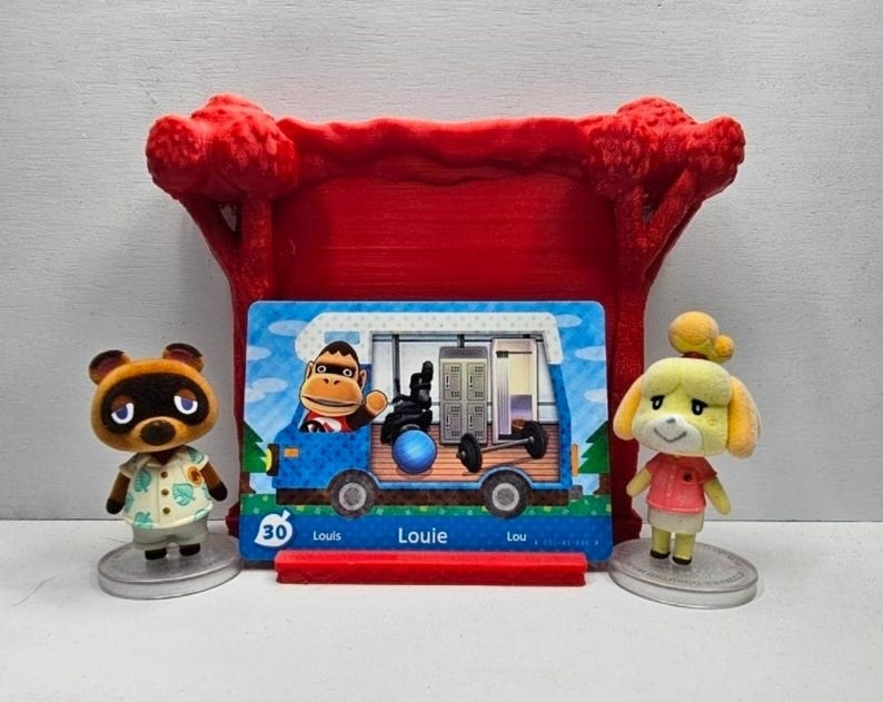 May include: A red plastic display stand holding a rectangular Animal Crossing amiibo card featuring the character Louie in a blue camper van. The card shows the number 30 and the name Louie. Two small character figurines stand on either side.